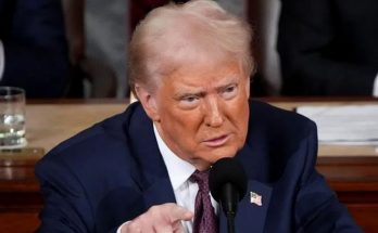 Donald Trump speaking into a microphone, wearing a dark blue suit, white shirt, and