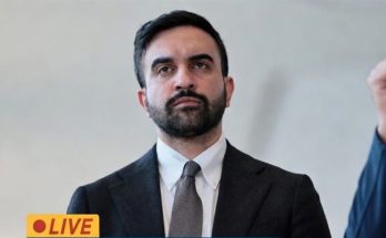 A man with dark hair and a beard wearing a dark suit, white shirt, and gray tie, with a