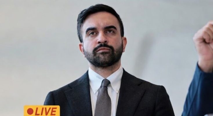 A man with dark hair and a beard wearing a dark suit, white shirt, and gray tie, with a