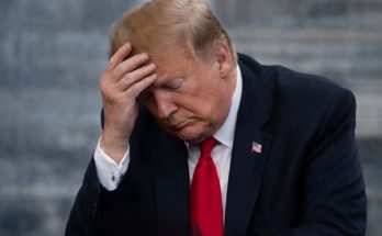 Donald Trump in a dark suit and red tie, holding his hand to his forehead with eyes closed