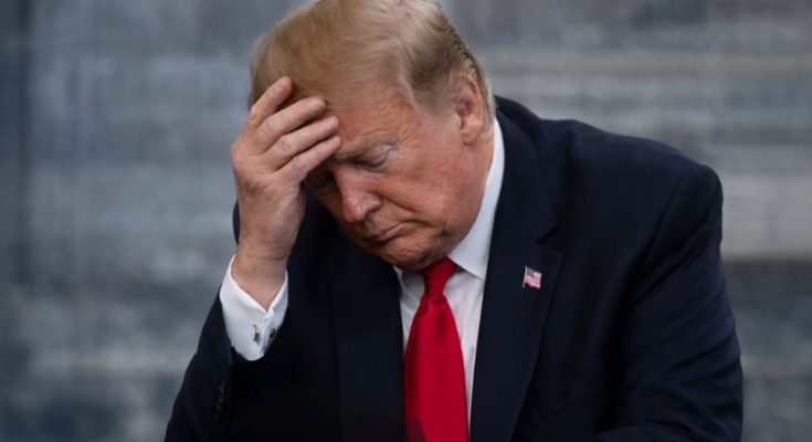 Donald Trump in a dark suit and red tie, holding his hand to his forehead with eyes closed