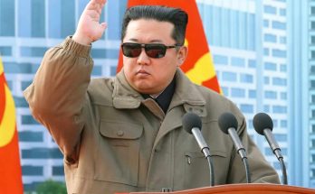 kim Jong Un wearing sunglasses and olive jumpsuit salutes at wooden podium with Workers