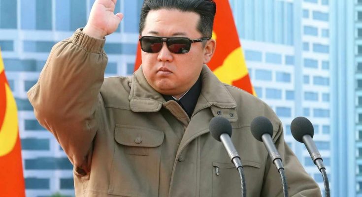 kim Jong Un wearing sunglasses and olive jumpsuit salutes at wooden podium with Workers