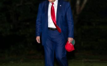 man in blue suit with red tie holding cap outdoors at night