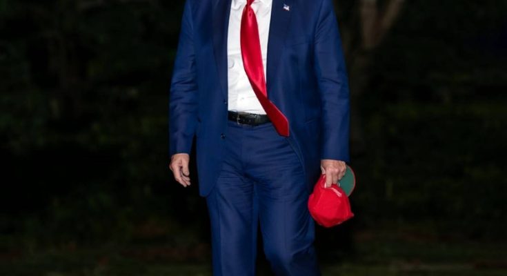 man in blue suit with red tie holding cap outdoors at night